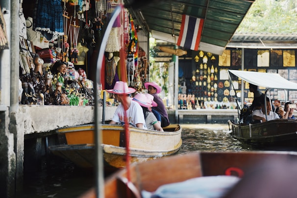 A bustling floating market scene with tourists wearing pink hats on a boat. Various souvenirs and colorful decorations line the walls of the market stalls. Another small boat is seen nearby, with people taking photos and observing the surroundings. The atmosphere is lively and vibrant.