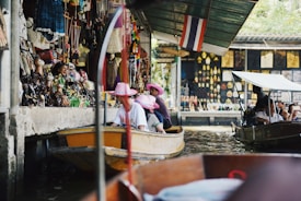 A bustling floating market scene with tourists wearing pink hats on a boat. Various souvenirs and colorful decorations line the walls of the market stalls. Another small boat is seen nearby, with people taking photos and observing the surroundings. The atmosphere is lively and vibrant.