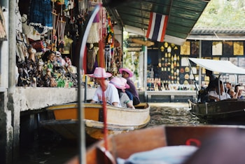 A bustling floating market scene with tourists wearing pink hats on a boat. Various souvenirs and colorful decorations line the walls of the market stalls. Another small boat is seen nearby, with people taking photos and observing the surroundings. The atmosphere is lively and vibrant.