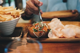 A table with a bowl of crispy fries and a tray containing fried snacks and a dish garnished with colorful herbs and vegetables. A hand is holding a utensil, suggesting preparation or serving.