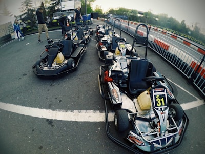 A group of go-karts parked on an outdoor track, surrounded by metal railings. The track has red and white barriers. People are visible in the background near the track, with trees and greenery surrounding the area.