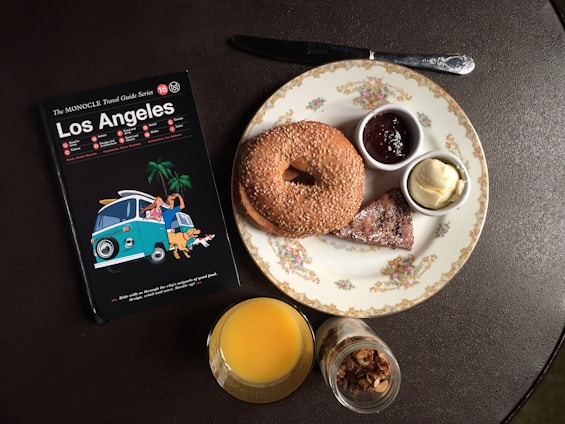 A table setting includes a travel guidebook for Los Angeles, a plate with a sesame bagel, small bowls of butter and jam, a croissant piece, a glass of orange juice, and a small jar of granola.