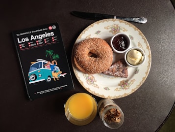 A table setting includes a travel guidebook for Los Angeles, a plate with a sesame bagel, small bowls of butter and jam, a croissant piece, a glass of orange juice, and a small jar of granola.