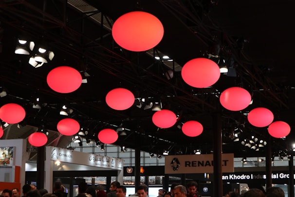 A ceiling with multiple large red spherical lights creating an ambient glow. Below, a crowd of people is visible, seemingly attending an event or exhibition. Numerous signs and booths, including those of automotive brands like Citroën and Renault, are present, indicating this might be an auto show or similar gathering.