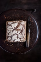 A loaf of rustic sourdough bread with a crusty, textured surface sits on a dark wooden table, accompanied by a vintage-style knife with a wooden handle.