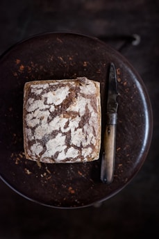 A loaf of rustic sourdough bread with a crusty, textured surface sits on a dark wooden table, accompanied by a vintage-style knife with a wooden handle.