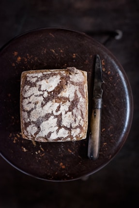 A loaf of rustic sourdough bread with a crusty, textured surface sits on a dark wooden table, accompanied by a vintage-style knife with a wooden handle.