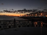 A seaside view at dusk with a silhouette of an ornate railing in the foreground. The ocean displays gentle waves and a row of illuminated buildings along a promenade stretches into the distance. The sky transitions from deep blue at the top to warm orange near the horizon, and scattered clouds add texture to the scene.