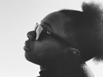 A monochrome profile portrait of a person wearing sunglasses. The person's hair is styled in a natural afro puff. The lighting creates contrast, highlighting the contours of the face.