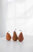 Three pears are neatly arranged on a white surface with a softly diffused window light in the background. The composition emphasizes simplicity and natural aesthetics.