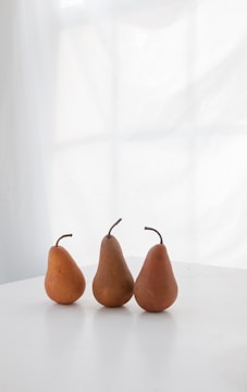 Three pears are neatly arranged on a white surface with a softly diffused window light in the background. The composition emphasizes simplicity and natural aesthetics.