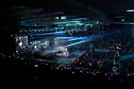 A large indoor arena filled with an audience watching a live performance. The stage features large screens displaying a performer and graphics, with bright spotlights and ambient lighting creating a dynamic atmosphere. The crowd is holding glow sticks, adding vibrant colors to the scene.