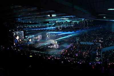 A large indoor arena filled with an audience watching a live performance. The stage features large screens displaying a performer and graphics, with bright spotlights and ambient lighting creating a dynamic atmosphere. The crowd is holding glow sticks, adding vibrant colors to the scene.