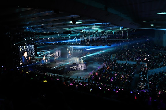 A large indoor arena filled with an audience watching a live performance. The stage features large screens displaying a performer and graphics, with bright spotlights and ambient lighting creating a dynamic atmosphere. The crowd is holding glow sticks, adding vibrant colors to the scene.