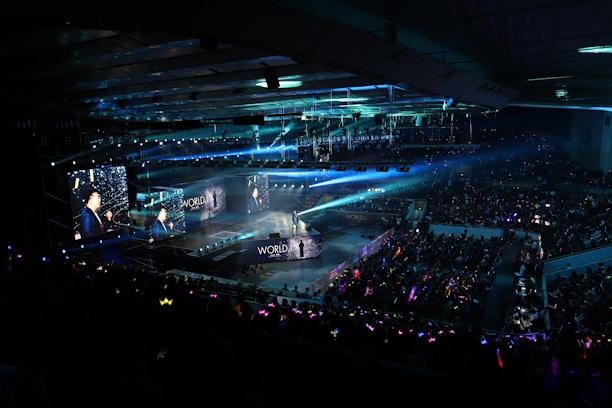 A large indoor arena filled with an audience watching a live performance. The stage features large screens displaying a performer and graphics, with bright spotlights and ambient lighting creating a dynamic atmosphere. The crowd is holding glow sticks, adding vibrant colors to the scene.