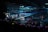 A large indoor arena filled with an audience watching a live performance. The stage features large screens displaying a performer and graphics, with bright spotlights and ambient lighting creating a dynamic atmosphere. The crowd is holding glow sticks, adding vibrant colors to the scene.