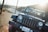 A black Jeep is parked on a dirt roadside with a mountainous landscape in the background. The sun is shining brightly, casting light flares and creating a warm glow in the image. Telephone poles line the road that curves into the distance, emphasizing a sense of open road and adventure.