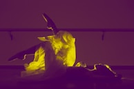 A ballet dancer is lying on the floor with one leg raised, showcasing a glowing yellow tutu against a purple background. The scene is backlit, emphasizing the delicate texture of the fabric and the graceful pose.