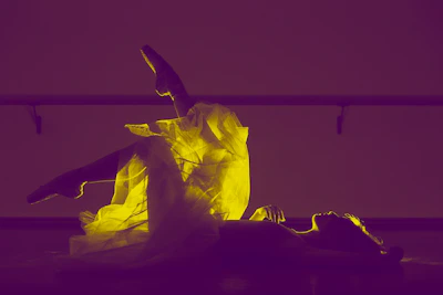A ballet dancer is lying on the floor with one leg raised, showcasing a glowing yellow tutu against a purple background. The scene is backlit, emphasizing the delicate texture of the fabric and the graceful pose.