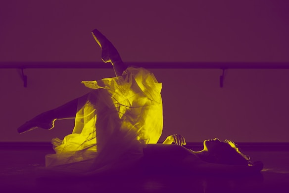 A ballet dancer is lying on the floor with one leg raised, showcasing a glowing yellow tutu against a purple background. The scene is backlit, emphasizing the delicate texture of the fabric and the graceful pose.
