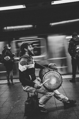 A street musician plays a drum with enthusiasm at a subway station. In the background, a train is captured in motion, creating a blurred effect. The musician is seated and smiling, wearing casual attire including a cap and jacket. People stand or walk by, immersed in their own activities.