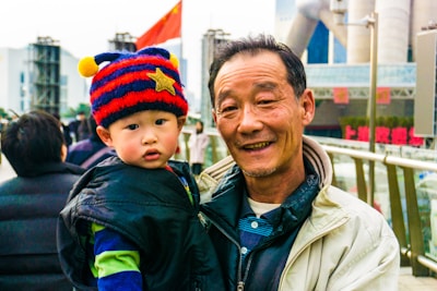 An elderly man holding a young child on a bustling urban street. The child is wearing a colorful hat with stripes and a star, while the man is dressed in a light jacket. Behind them, modern buildings and a red flag are visible.