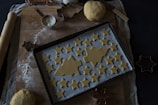 A wooden surface is covered with a cloth and lightly dusted with flour. On it rests a baking tray lined with parchment paper, topped with unbaked star and tree-shaped cookies. A ball of dough, a rolling pin, a metal measuring cup filled with flour, and various cookie cutters are scattered around. The lighting is dim, creating a cozy atmosphere.