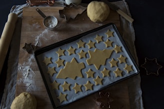 A wooden surface is covered with a cloth and lightly dusted with flour. On it rests a baking tray lined with parchment paper, topped with unbaked star and tree-shaped cookies. A ball of dough, a rolling pin, a metal measuring cup filled with flour, and various cookie cutters are scattered around. The lighting is dim, creating a cozy atmosphere.
