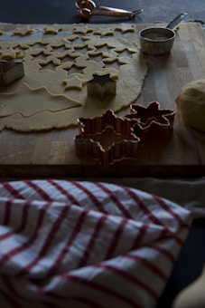 A wooden surface is covered with rolled-out dough, with numerous star-shaped cutouts. Metal cookie cutters are scattered around, and a measuring cup with flour is to the side. A striped red and white kitchen towel is partially visible, adding a cozy touch to the scene.