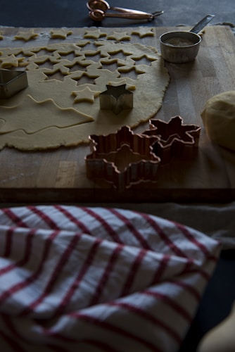 A wooden surface is covered with rolled-out dough, with numerous star-shaped cutouts. Metal cookie cutters are scattered around, and a measuring cup with flour is to the side. A striped red and white kitchen towel is partially visible, adding a cozy touch to the scene.