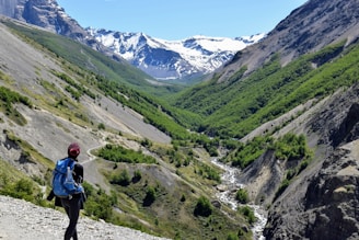 A hiker with a blue backpack stands on a rocky path overlooking a vast valley surrounded by green hills and towering snow-capped mountains. A small winding river cuts through the landscape, and the sky is clear and blue.