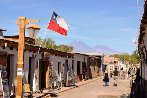 A street scene in a small town with rustic, low-rise buildings lining both sides. A flag can be seen waving on a pole, and there are signs and posters on the walls. Several people are walking down the unpaved street. In the background, distant mountains under a clear blue sky are visible.