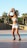 A woman prepares to shoot a basketball, standing on an outdoor court with palm trees and a stone wall in the background. She is wearing a white crop top, denim shorts, and black sneakers. The sun casts long shadows, indicating it is late afternoon.