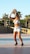 A woman prepares to shoot a basketball, standing on an outdoor court with palm trees and a stone wall in the background. She is wearing a white crop top, denim shorts, and black sneakers. The sun casts long shadows, indicating it is late afternoon.