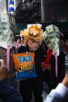 A person is wearing a mask resembling a prominent public figure, adorned with a makeshift crown and straw-like hair. They are flanked by two individuals with large paper mache representations of dollar bills on their heads. The center figure holds a bag labeled 'Tax Cheeto Pres' and a large pair of red scissors.