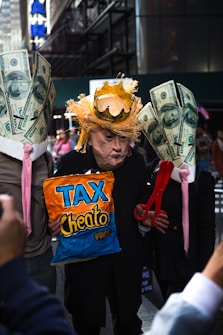 A person is wearing a mask resembling a prominent public figure, adorned with a makeshift crown and straw-like hair. They are flanked by two individuals with large paper mache representations of dollar bills on their heads. The center figure holds a bag labeled 'Tax Cheeto Pres' and a large pair of red scissors.