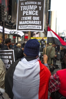 A protester is wrapped in a flag with the colors black, white, and red, holding a sign demanding the removal of the Trump and Pence regime. The setting appears to be an urban environment with other people nearby and buildings in the background.