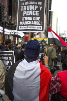 A protester is wrapped in a flag with the colors black, white, and red, holding a sign demanding the removal of the Trump and Pence regime. The setting appears to be an urban environment with other people nearby and buildings in the background.