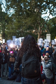 A person with long dark hair and a backpack stands before a large crowd holding protest signs. The gathering takes place outdoors in a park-like area with trees and some structures visible in the background. Many people are using cameras or phones to document the event.