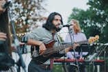 A musician with long hair plays a black electric bass guitar while singing into a microphone. He is dressed casually in a patterned shirt and tie. In the background, two other musicians are visible, one playing a keyboard. The scene is set outdoors with trees and string lights, suggesting a casual, perhaps festive, atmosphere.