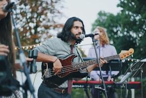 A musician with long hair plays a black electric bass guitar while singing into a microphone. He is dressed casually in a patterned shirt and tie. In the background, two other musicians are visible, one playing a keyboard. The scene is set outdoors with trees and string lights, suggesting a casual, perhaps festive, atmosphere.