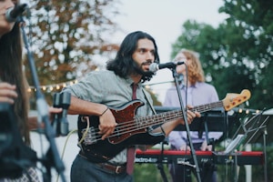 A musician with long hair plays a black electric bass guitar while singing into a microphone. He is dressed casually in a patterned shirt and tie. In the background, two other musicians are visible, one playing a keyboard. The scene is set outdoors with trees and string lights, suggesting a casual, perhaps festive, atmosphere.