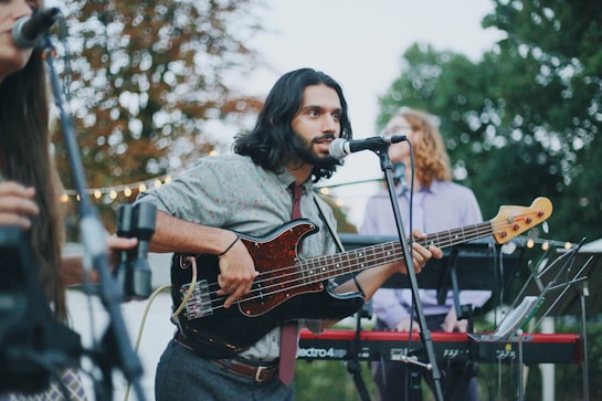 A musician with long hair plays a black electric bass guitar while singing into a microphone. He is dressed casually in a patterned shirt and tie. In the background, two other musicians are visible, one playing a keyboard. The scene is set outdoors with trees and string lights, suggesting a casual, perhaps festive, atmosphere.