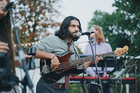 A musician with long hair plays a black electric bass guitar while singing into a microphone. He is dressed casually in a patterned shirt and tie. In the background, two other musicians are visible, one playing a keyboard. The scene is set outdoors with trees and string lights, suggesting a casual, perhaps festive, atmosphere.