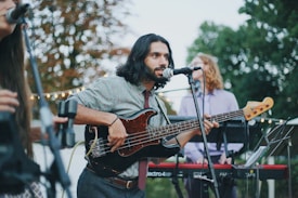 A musician with long hair plays a black electric bass guitar while singing into a microphone. He is dressed casually in a patterned shirt and tie. In the background, two other musicians are visible, one playing a keyboard. The scene is set outdoors with trees and string lights, suggesting a casual, perhaps festive, atmosphere.