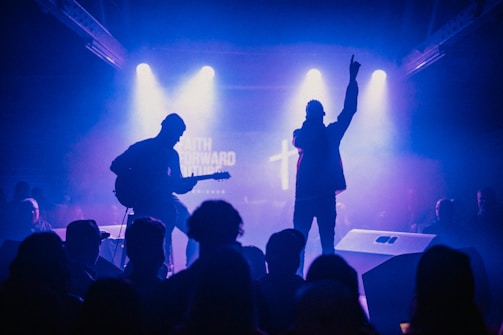 Silhouetted performers on stage in a dimly lit venue, with one person playing the guitar and another holding a microphone. The background is illuminated by blue and purple stage lights, creating a dramatic contrast.