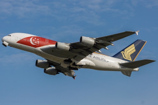 A large commercial airplane in flight with a prominent Singaporean flag design on its body. The aircraft features a blue and gold tail with a distinctive logo and is set against a backdrop of clear blue sky.