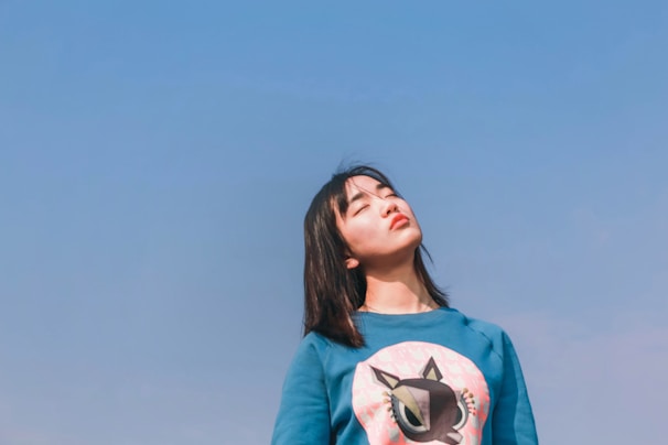 A person stands against a clear blue sky with eyes closed, appearing relaxed. The individual wears a blue sweatshirt featuring a cartoon graphic. The sun casts gentle light on their face, creating a serene and peaceful atmosphere.