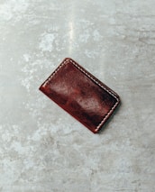 A small, rectangular brown leather wallet with visible stitching is placed on a textured, grey surface.