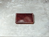 A dark brown leather wallet with white stitching along its edges is placed on a textured, gray surface. The wallet's surface appears glossy with some natural creases visible.